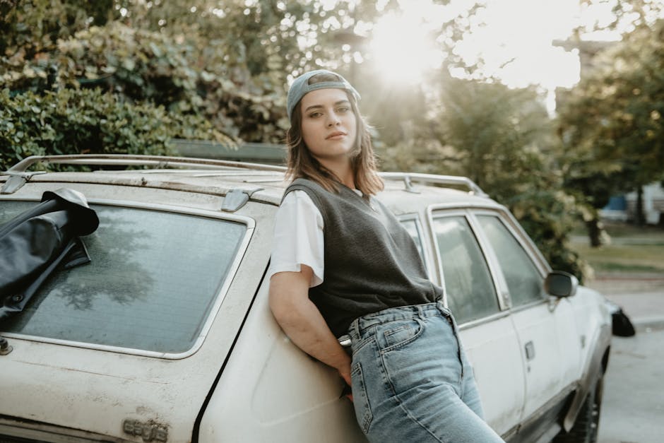 A young woman in casual attire leans against a vintage car in an outdoor setting with sunlight filtering through trees.