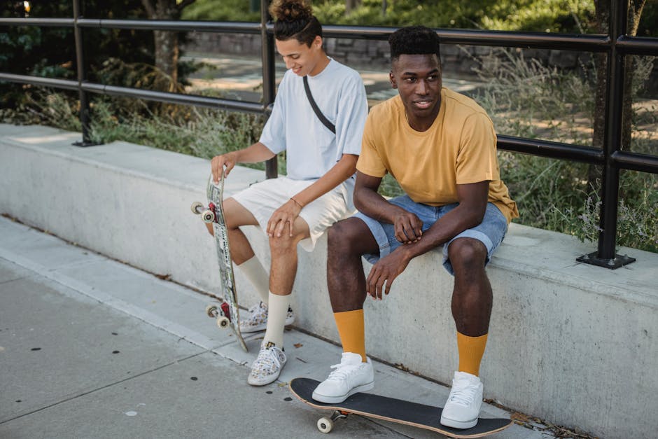 Full body young content multiracial friends with skateboards wearing casual summer clothes sitting on walkway in lush park