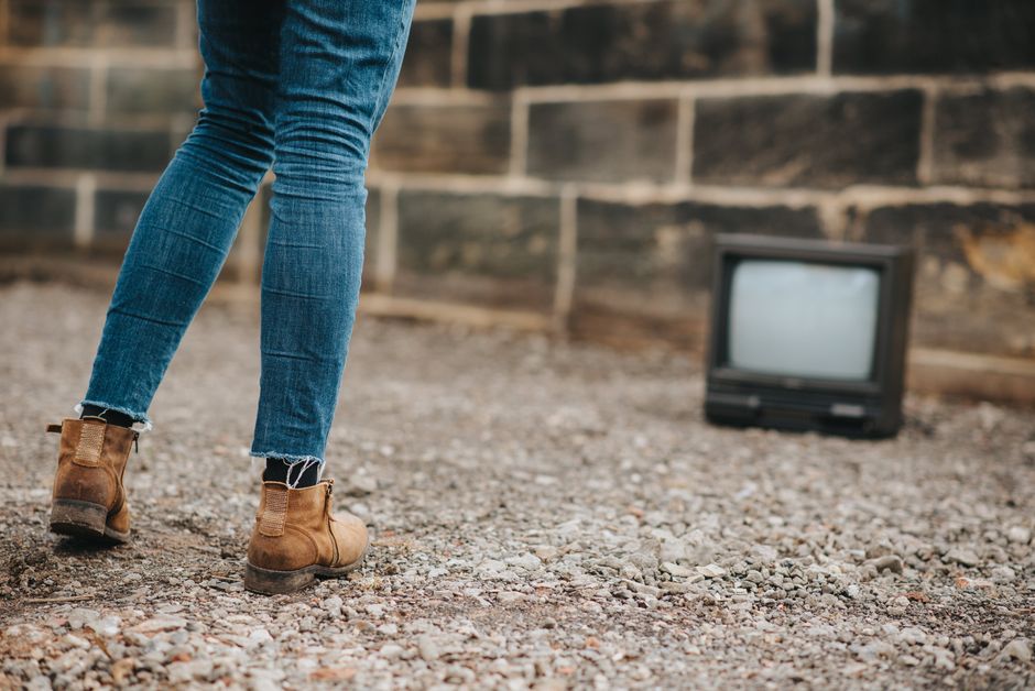 Back view of anonymous female in jeans standing on ground against stone wall and old fashioned television on blurred background