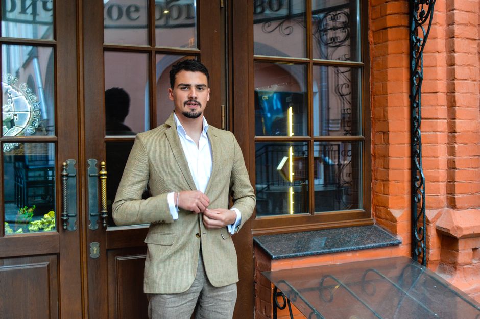 Confident bearded male in elegant suit standing on street near brick building with wooden doors on city while looking at camera