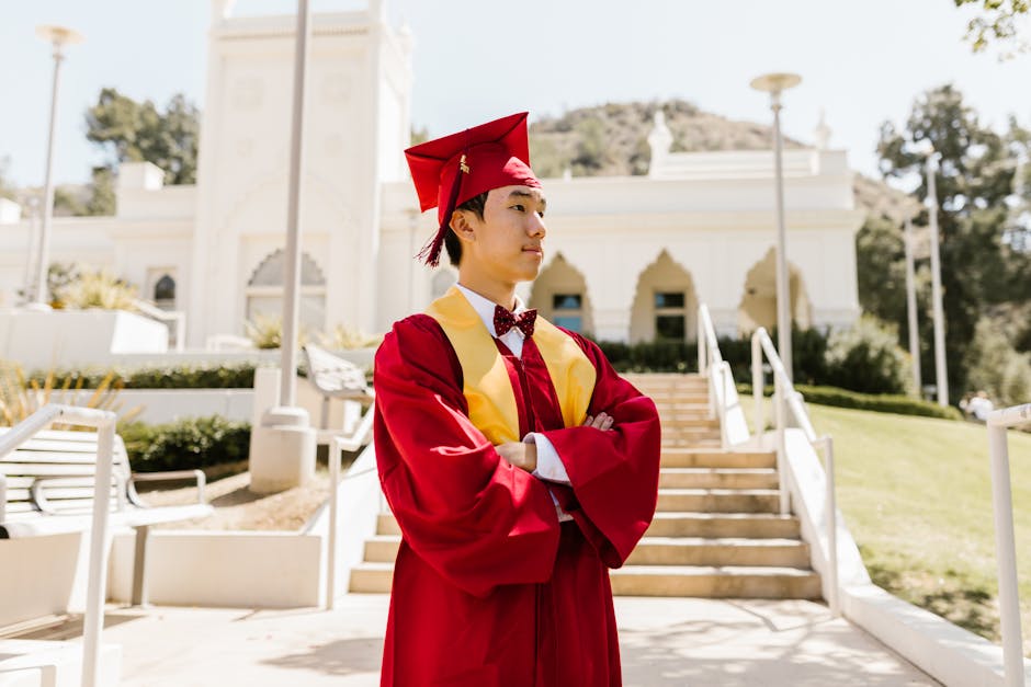 A proud graduate in a red gown confidently poses outdoors in front of a historical building.