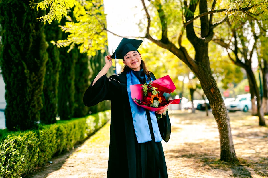 Young female graduate in cap and gown holding a bouquet in a sunny outdoor setting.