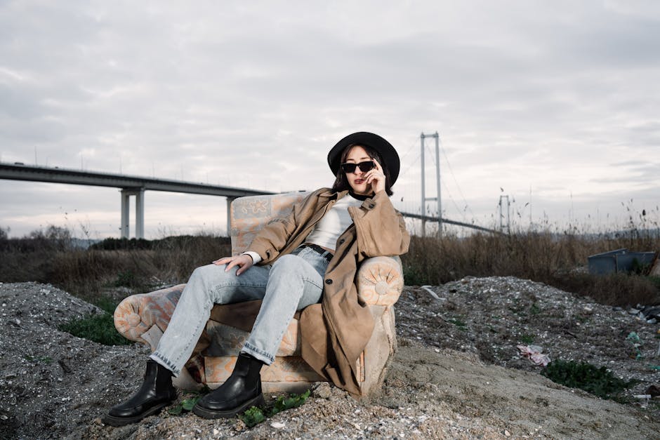 Stylish woman in a trench coat and hat lounging outdoors near a bridge, exuding a modern vibe.