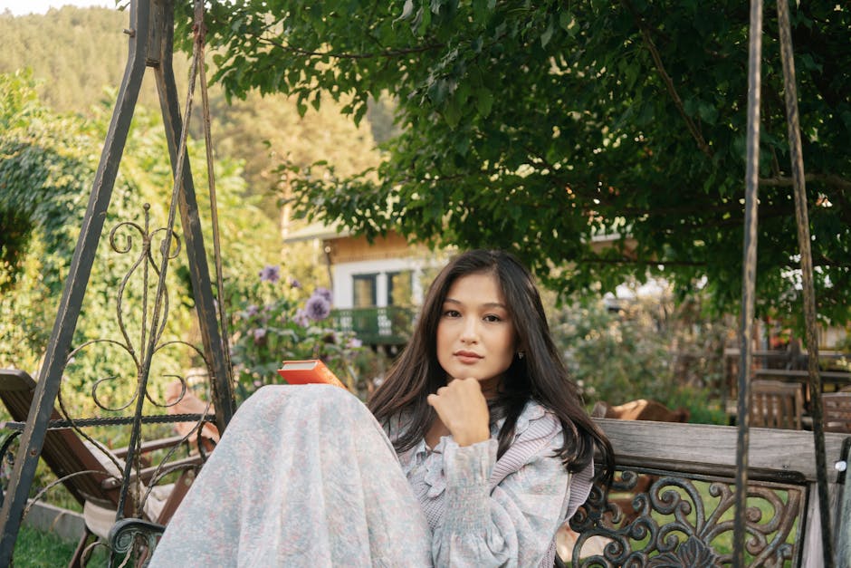 A young Asian woman sits on a swing in a garden, enjoying a book on a peaceful day.