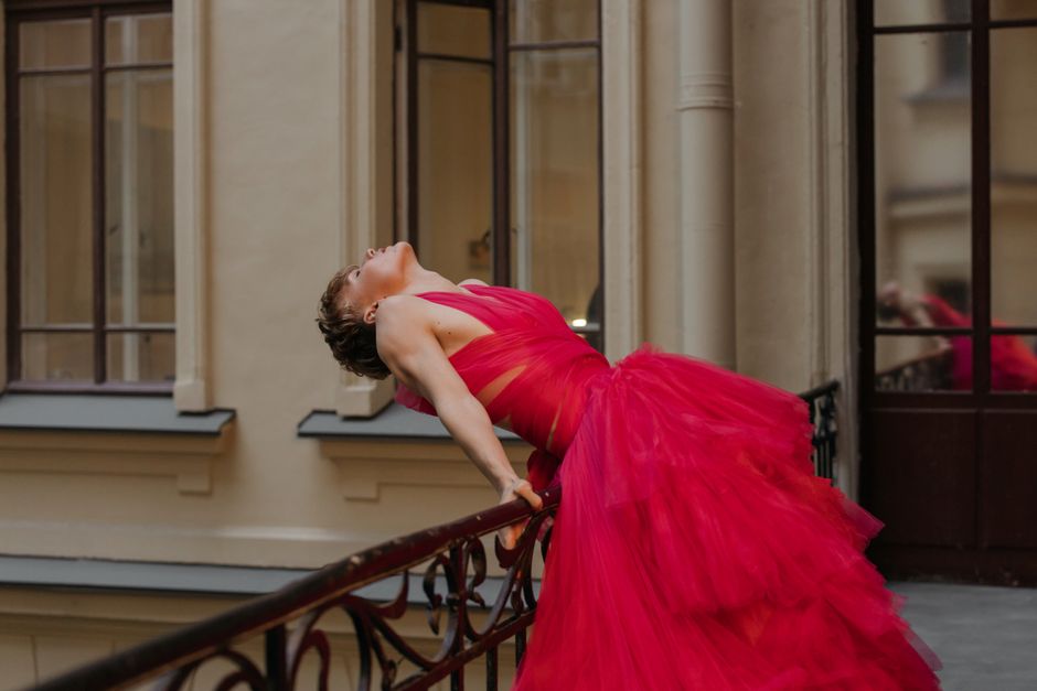 A woman in a striking red dress elegantly leans back on a metal balcony railing, exuding fashion and grace.