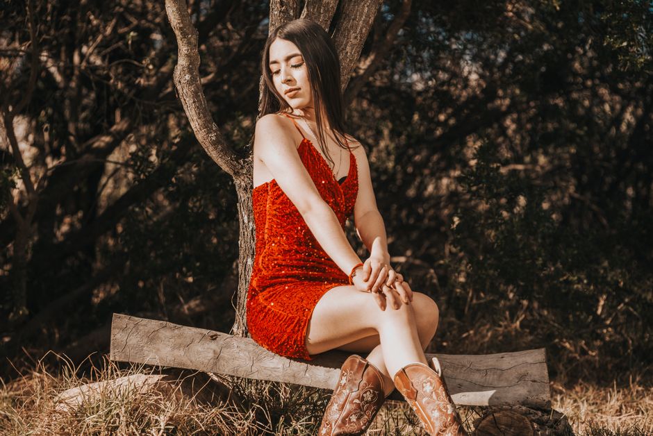 A woman in a red dress and cowboy boots sits on a wooden bench in a rural setting, showcasing fashion and elegance.
