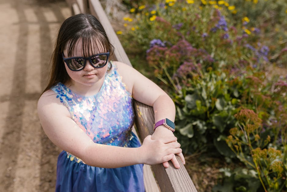 Young girl in sunglasses and sequin dress stands outside, surrounded by flowers.