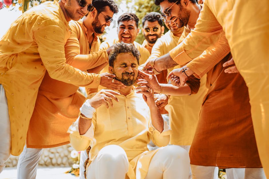 A joyful group celebrating a groom's haldi ceremony outdoors, wearing traditional attire.