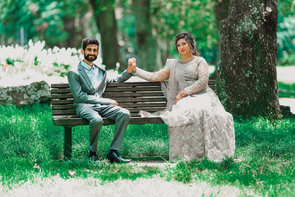 A couple in wedding attire holding hands on a park bench, surrounded by lush greenery.